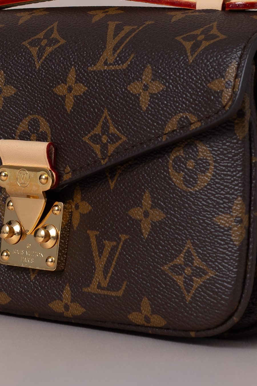 Close-up de uma bolsa: fecho metálico dourado com rebites, costura aparente em linha clara e vivo em couro natural.